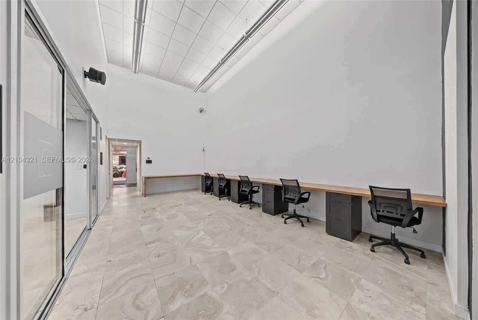 High-ceiling workspace featuring expansive stone-tiled flooring, extensive built-in wooden desk surfaces, dark cabinetry, and floor-to-ceiling glass paneling