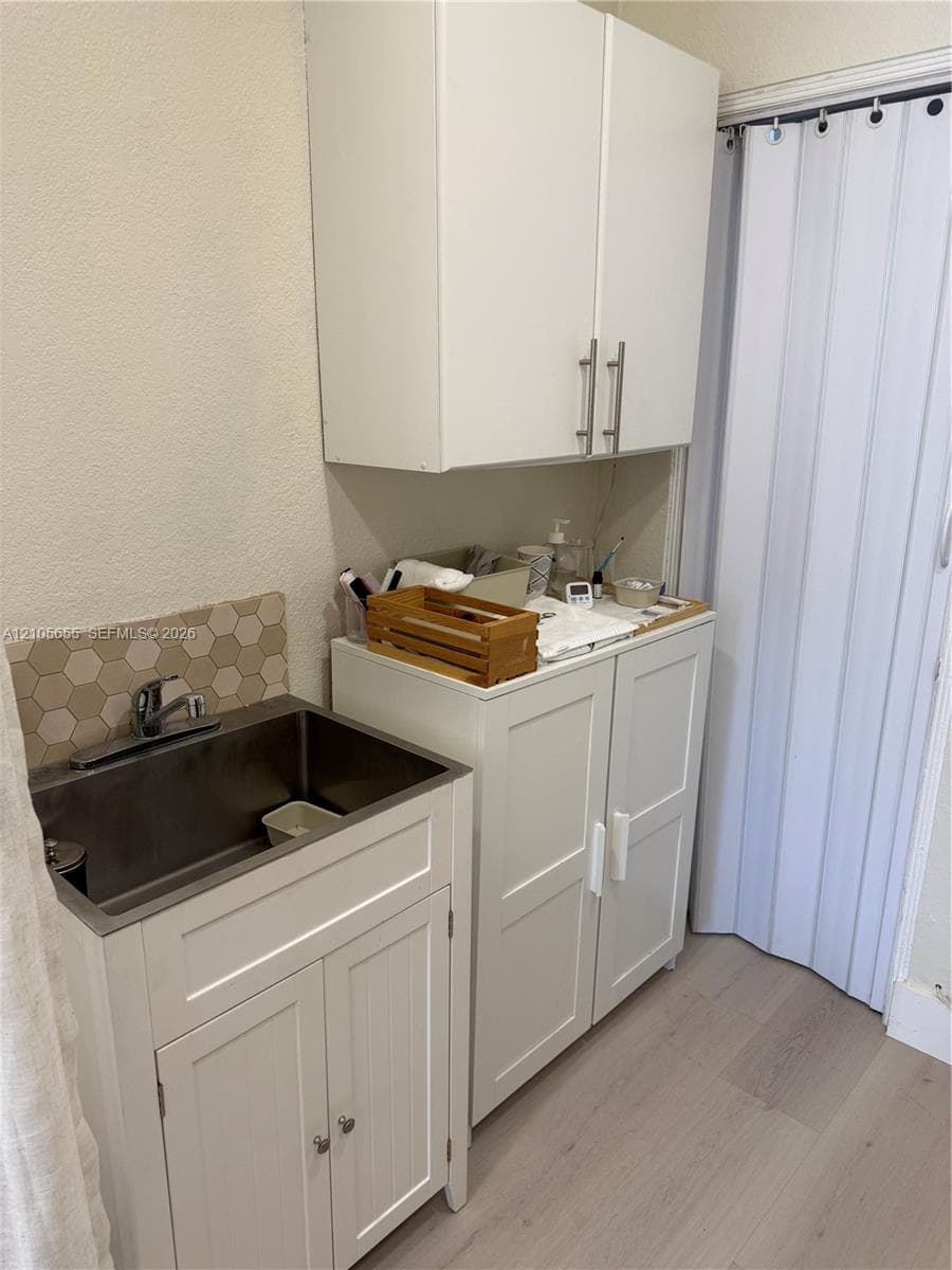 Utility area featuring white shaker-style cabinetry, a stainless steel basin sink, hexagonal tile backsplash, and light-toned wood-finish flooring