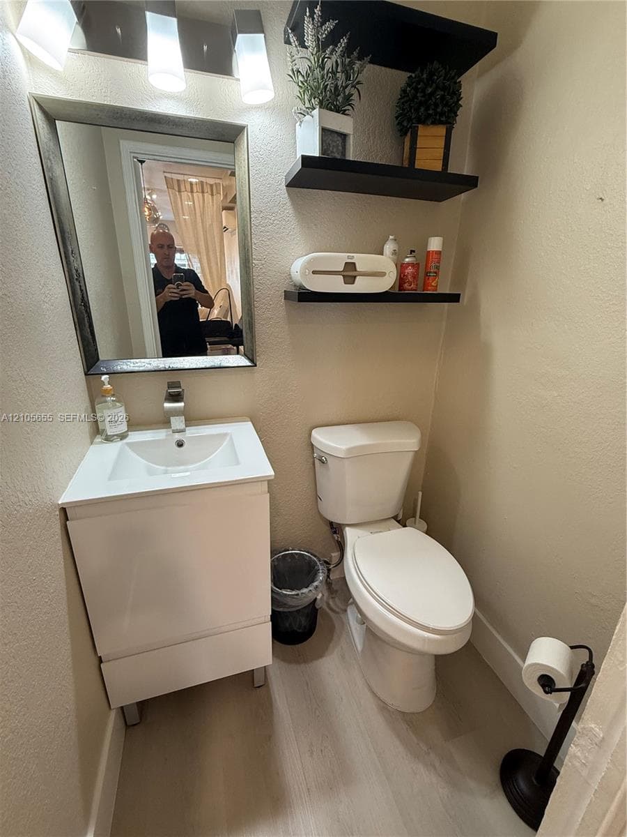 White pedestal-style vanity with integrated sink, rectangular wall-mounted mirror, light-colored wood-finish flooring, and black floating shelving