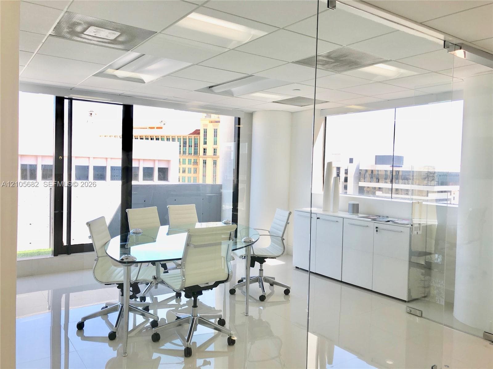 Glass-enclosed workspace featuring floor-to-ceiling windows, white tile flooring, drop ceiling with recessed lighting, and built-in white cabinetry