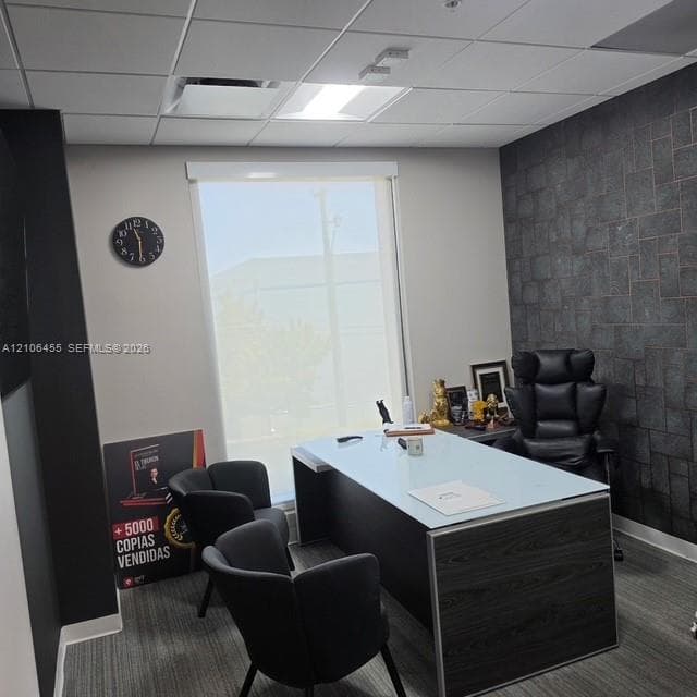 Office space featuring a textured dark stone accent wall, neutral wall tones, grid-pattern ceiling tiles, and window with a roller shade