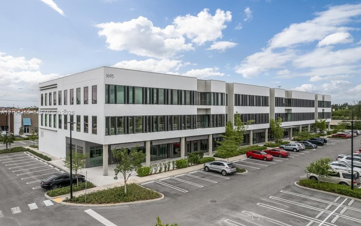 Multi-story commercial building featuring a white facade, extensive glass window panels, a covered ground-level walkway with support columns, and an adjacent asphalt parking lot