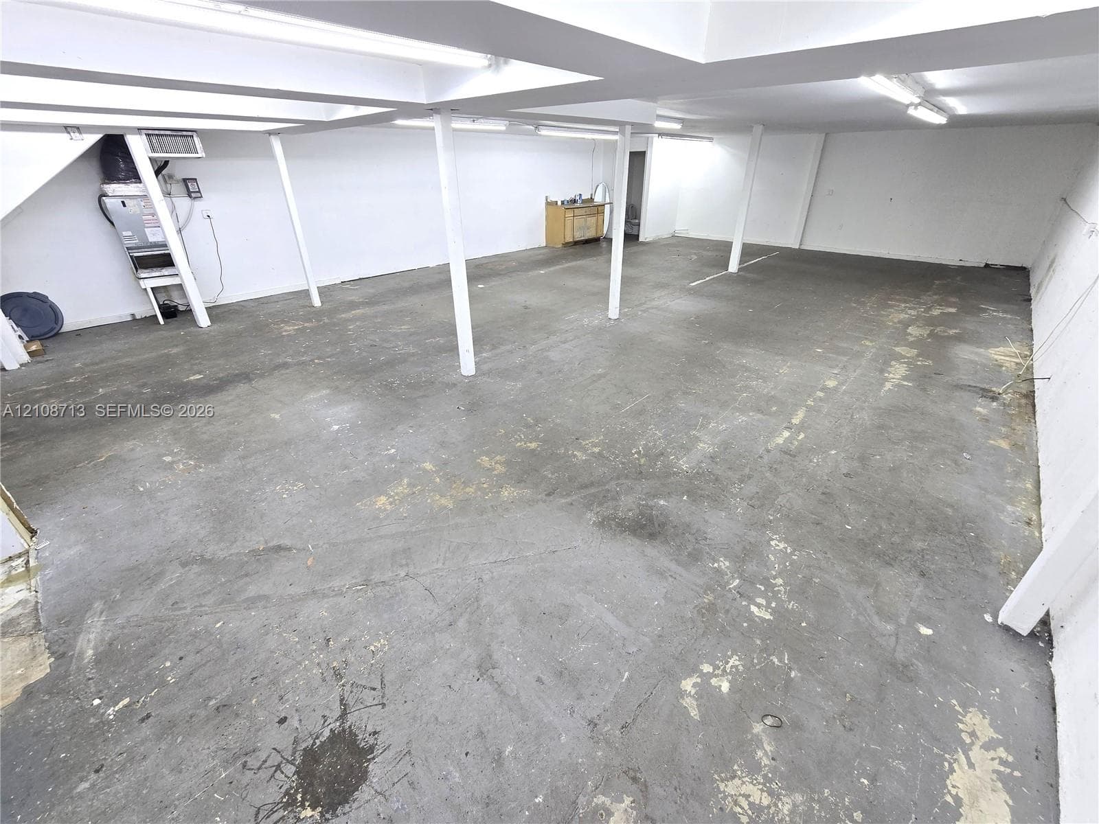 Expansive open-concept utility area featuring concrete flooring, structural support pillars, recessed ceiling lighting, and a neutral wall finish