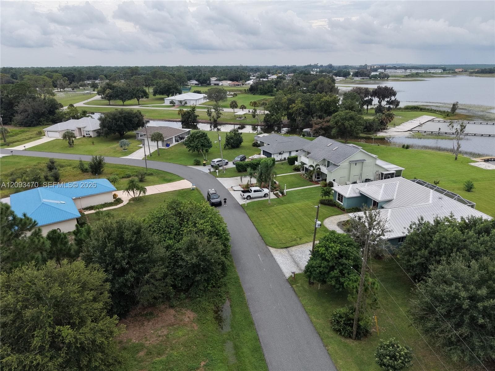Aerial perspective of a residential neighborhood featuring homes with metal roofs, asphalt-paved streets, mature trees, open green space, and proximity to a large body of water