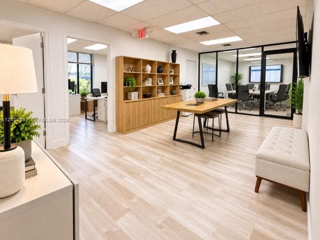 Open-concept office space featuring light wood-finish flooring, built-in floor-to-ceiling wooden shelving, glass-enclosed partitioned office space, and recessed drop-ceiling lighting
