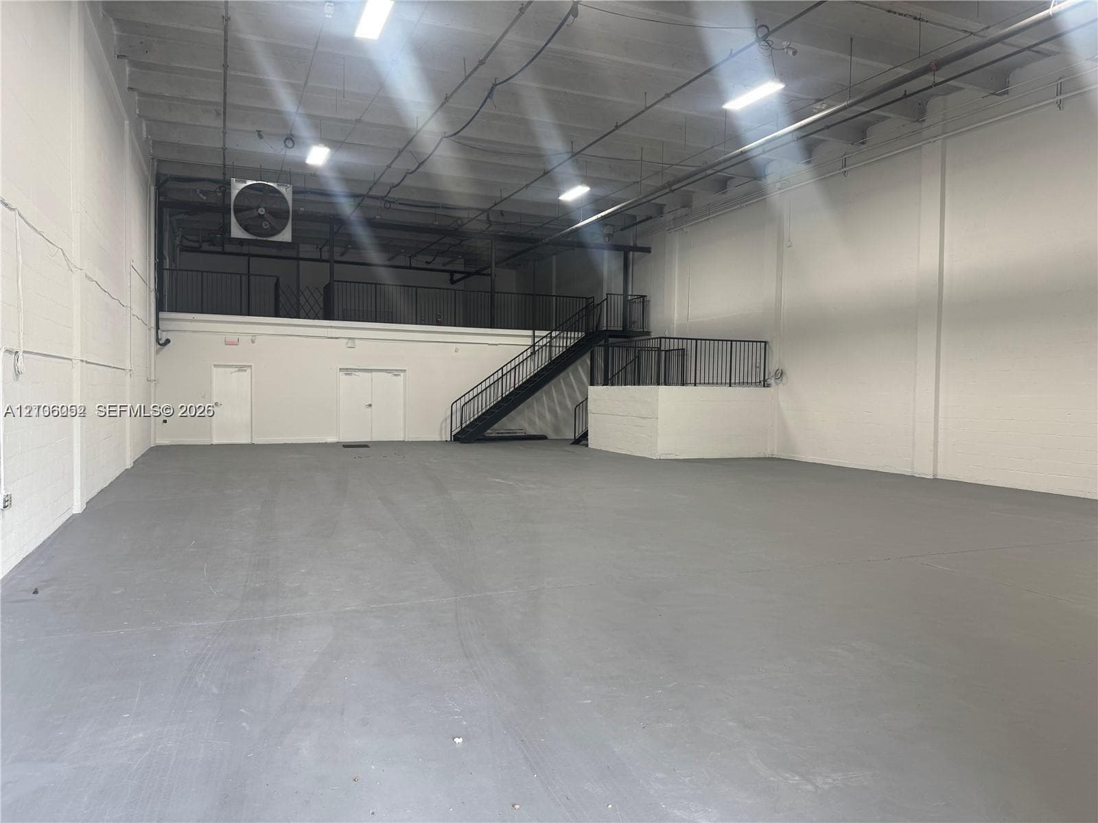 Open-concept industrial warehouse space featuring high ceilings, painted masonry walls, durable floor surfacing, mezzanine level with metal railing, and staircase access